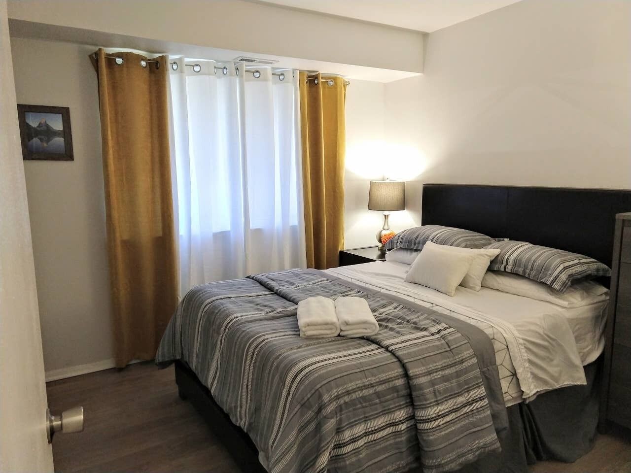 A hotel room with a bed, curtains, a nightstand with a lamp, and a picture on the wall.