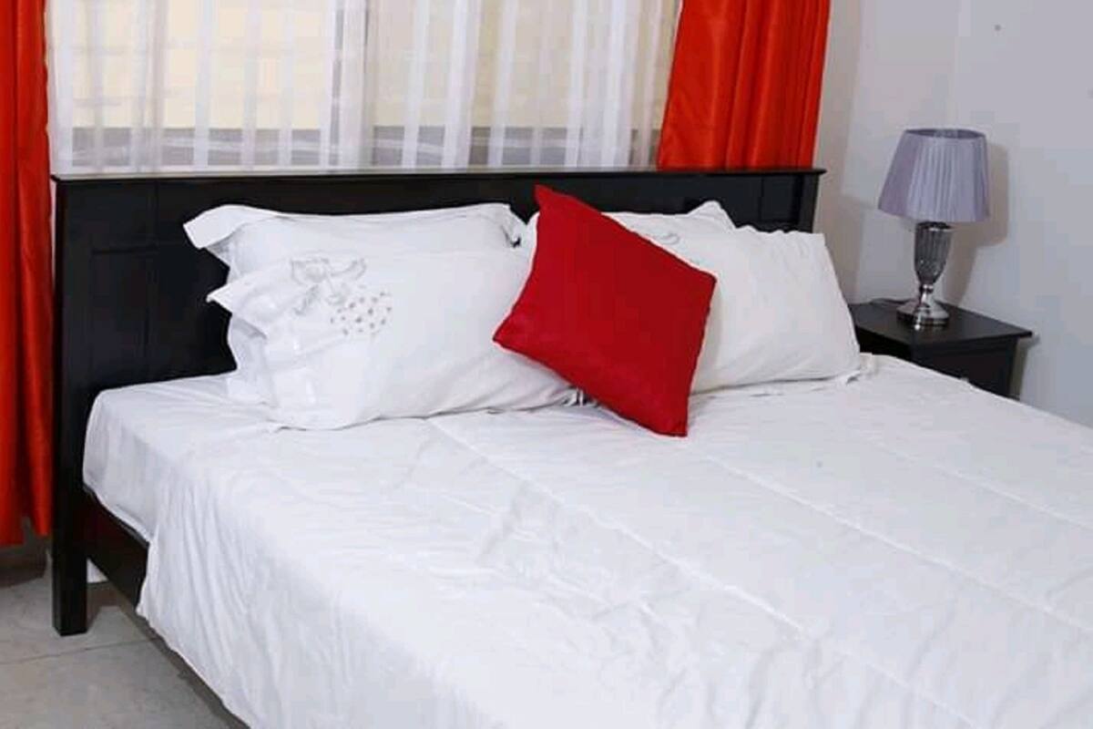 A bed with white bedding and a red pillow, a nightstand with a lamp, and red curtains.