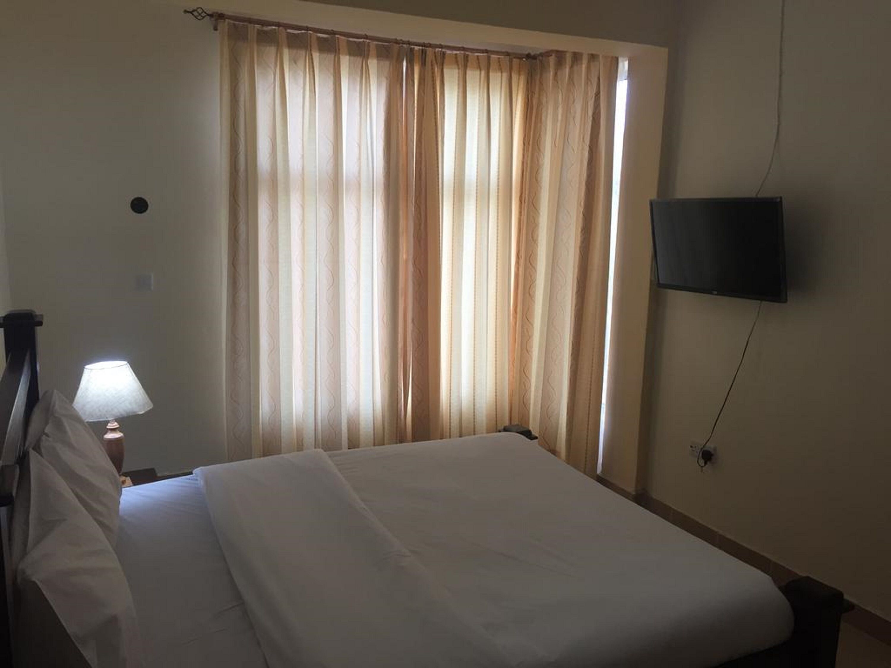 Habitación de hotel con cama, lámpara, televisor montado en la pared y ventana con cortinas.