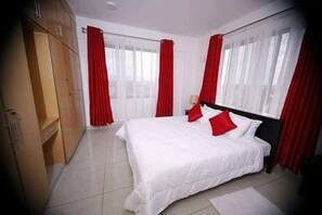 A bedroom with a large bed, red curtains, a wardrobe, and a window. - Milimani Hotel (Kakamega)