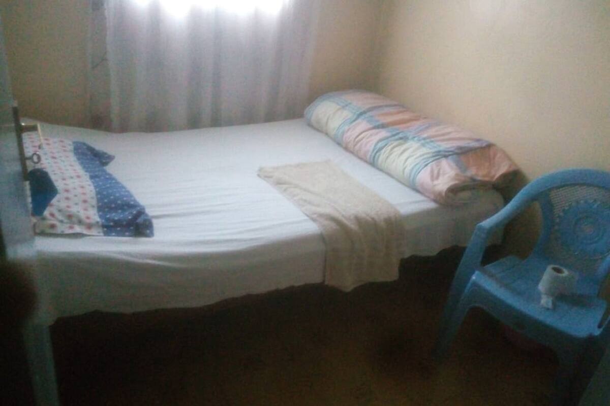 Quarto pequeno e simples com uma cama de solteiro, uma cadeira e uma janela com cortinas.