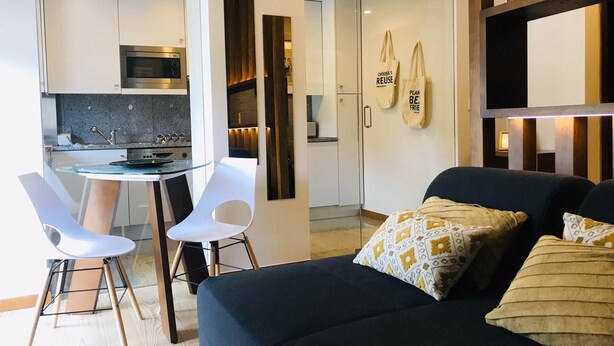 A modern living room with a black sofa, white chairs, and a glass coffee table. A kitchen with white cabinets and a microwave is visible in the background. - Flat Casino Beach Figueira da Foz - Moderno & Ótima Localização! (Figueira da Foz)