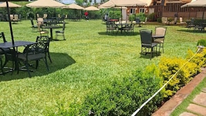 An outdoor patio area with tables and chairs under umbrellas, surrounded by well-maintained grass and hedges. - Sailors Hotel (Mlolongo)
