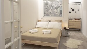 A bedroom with a bed, a ceiling fan, a window with blinds, a wall-mounted air conditioner, and a small bedside table with a lamp.