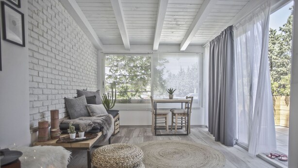 A cozy room with a white brick wall, a wooden ceiling, a sofa with cushions, a small table with chairs, a potted plant, and a large window with curtains.