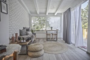 A cozy room with a white brick wall, a wooden ceiling, a sofa with cushions, a small table with chairs, a potted plant, and a large window with curtains. - Little Suite - Scala dei Turchi (Porto Empedocle)