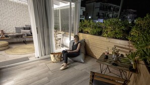 A modern living room with a sofa, a coffee table, and a dining table. There is a sliding glass door leading to a balcony with plants.