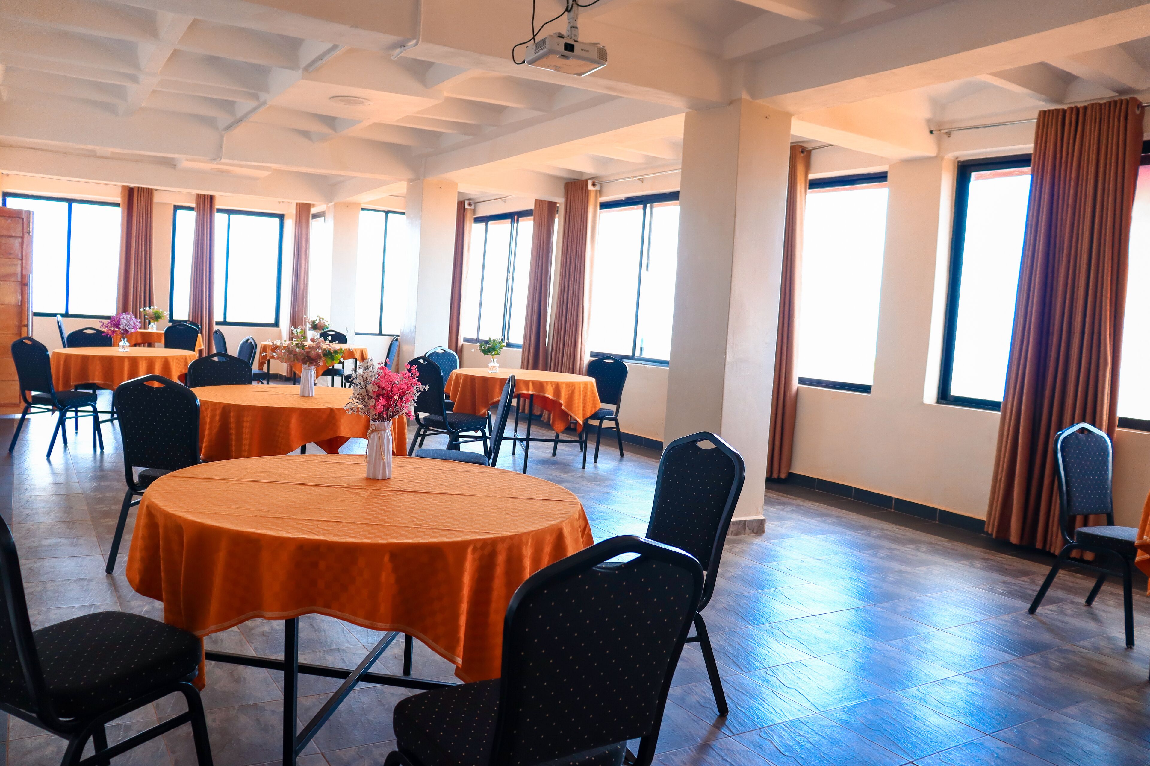 A conference room with multiple tables and chairs, large windows with curtains, and a tiled floor.