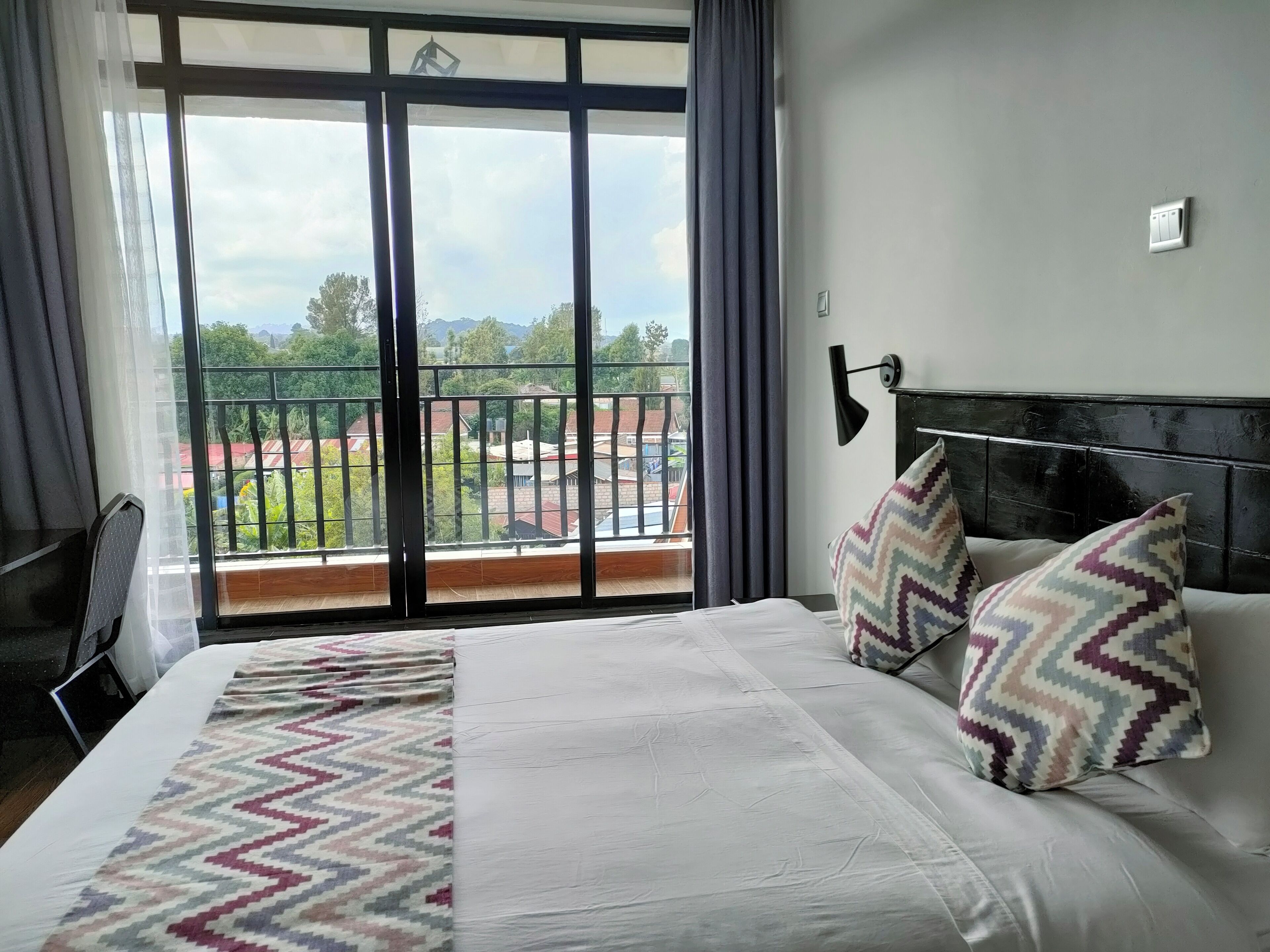 Una habitación de hotel con una cama grande, vistas a un balcón con barandilla y una mesita de noche con lámpara.