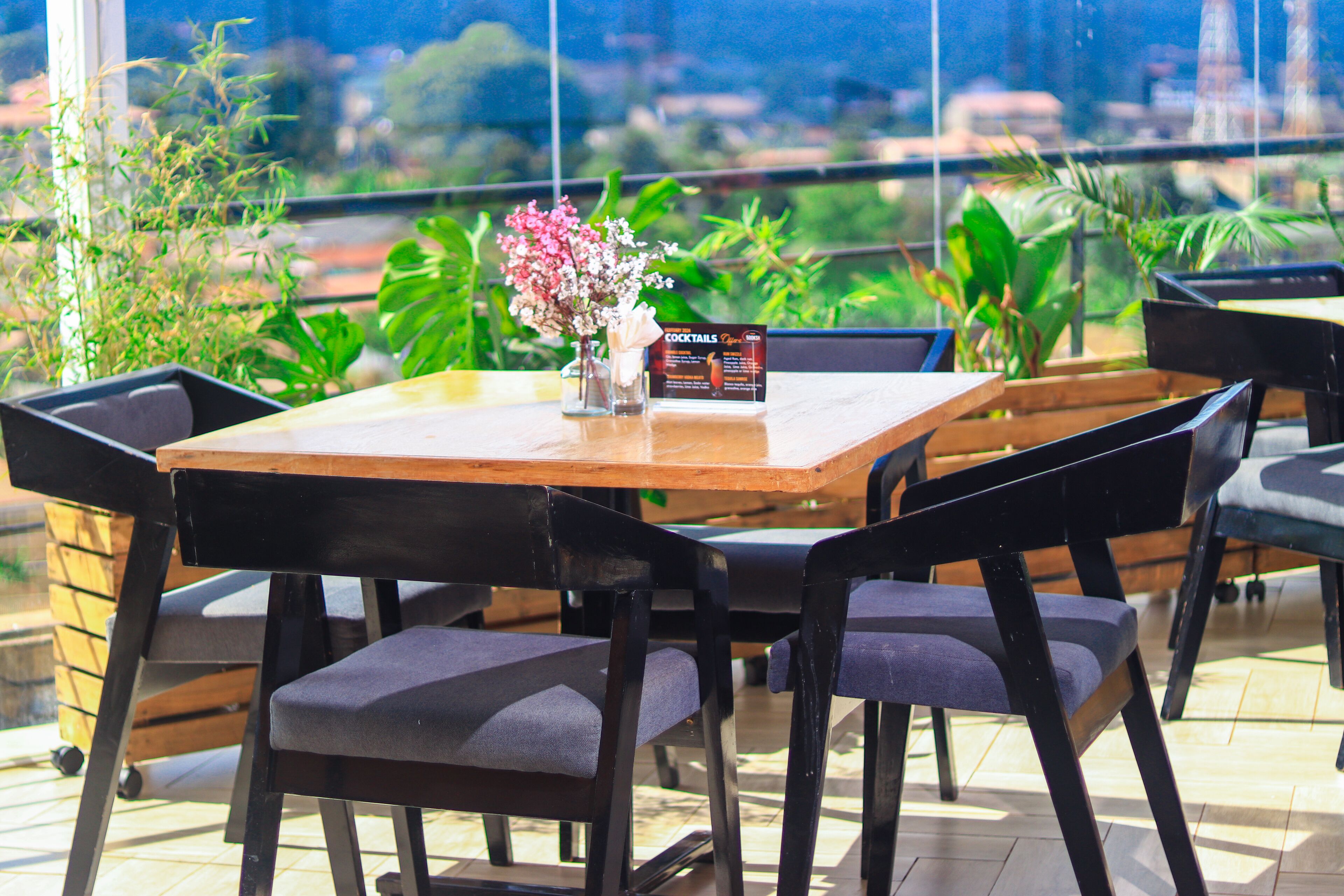 A table with chairs, a vase with flowers, and a menu sign in a restaurant setting with a view of greenery and buildings.