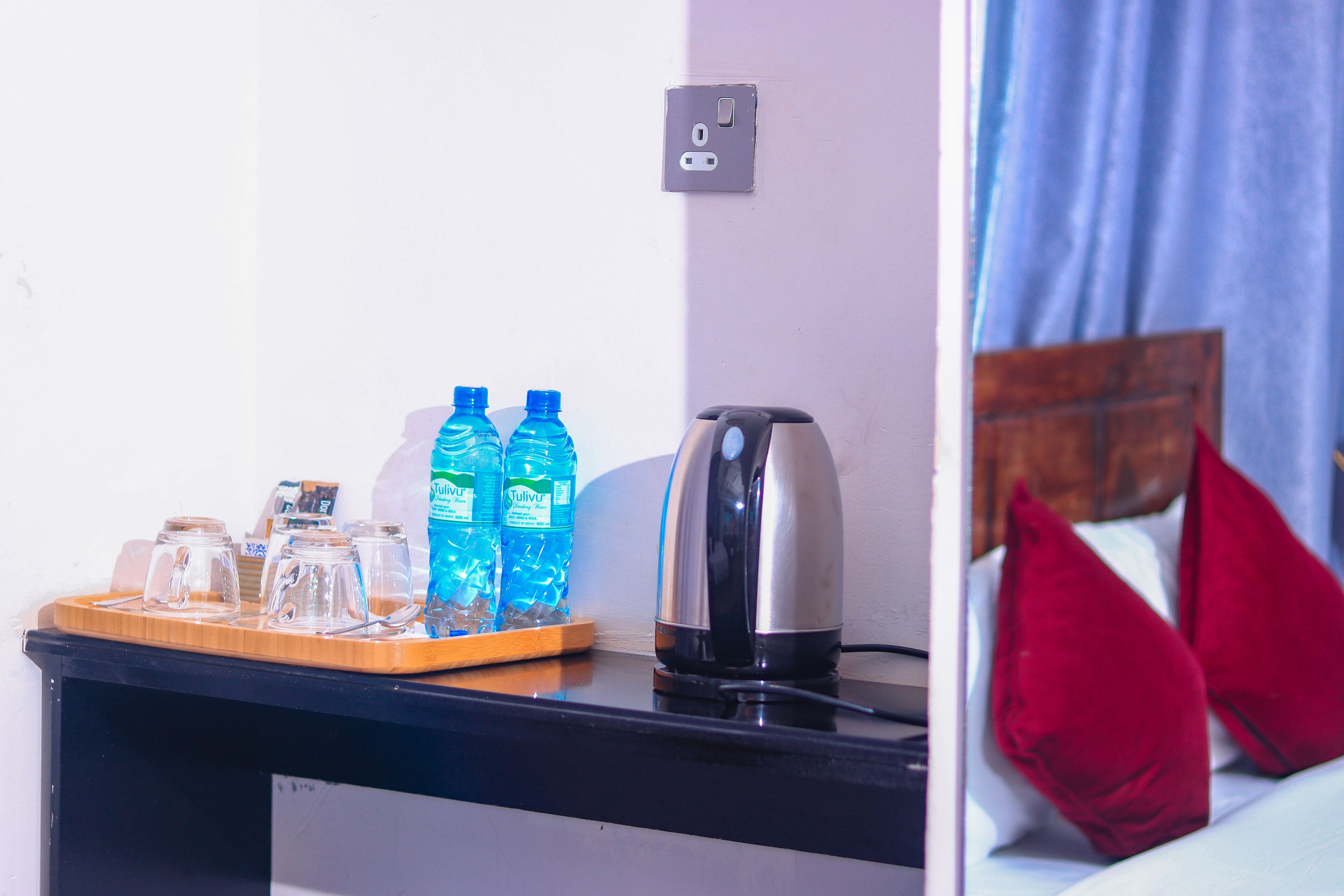 Une chambre d’hôtel avec un plateau en bois contenant des verres et des bouteilles d’eau, une bouilloire et une prise électrique au mur.