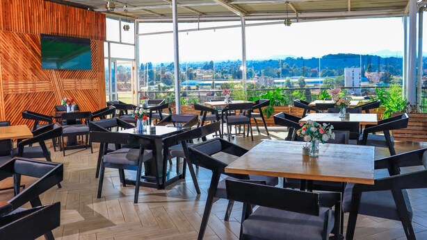 Un amplio espacio exterior para comer con varias mesas y sillas, ventanas grandes que ofrecen vistas a la ciudad y las colinas, y una pared de madera con una pantalla montada.