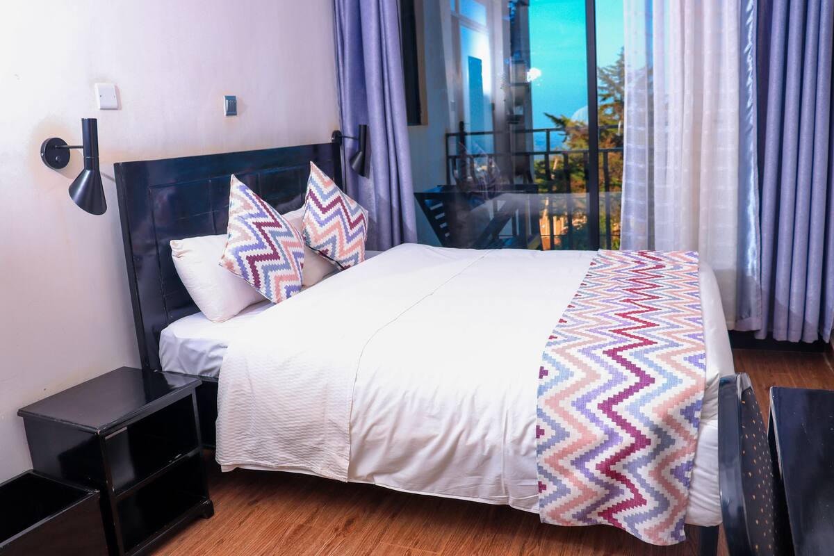 A neatly made bed with patterned pillows, a bedside table, and a view of the outdoors through a window with curtains.