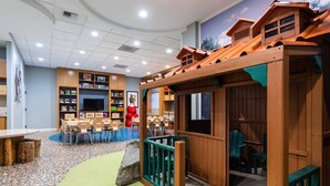 Zona infantil con una casita de madera, mesas, sillas y suelo alfombrado.