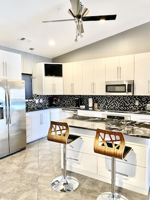 A modern kitchen with white cabinets, a stainless steel refrigerator, a microwave, and two bar stools with wooden seats.
