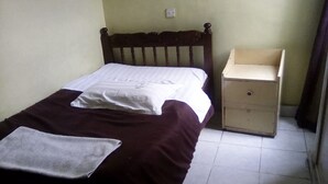 Una cama individual con cabecera de madera, una mesita de noche con cajón y una pared lisa con interruptor de luz.