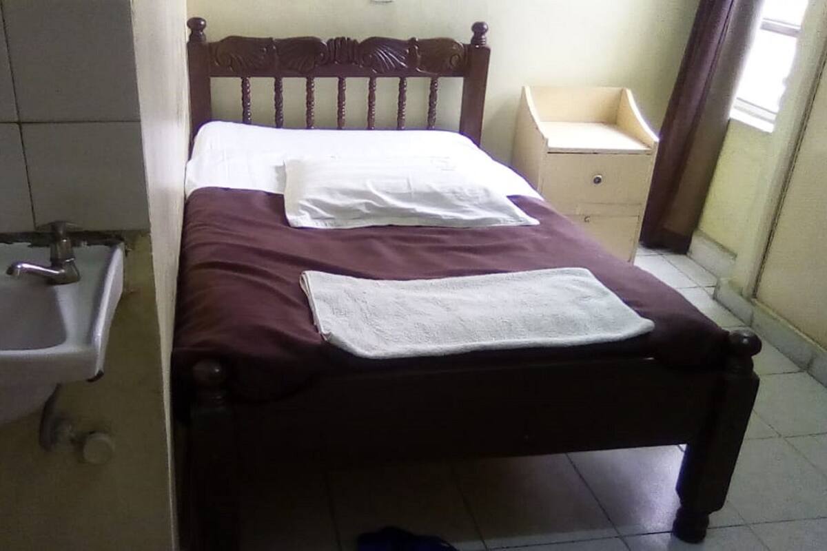 A single bed with a wooden headboard, a white pillow, and a white towel on the bed. There is a small bedside table with a drawer and a mirror. A white sink is visible to the left of the bed.