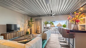 A modern living room with a large TV, a sofa, a dining area with chairs, and a view of the beach. - PM78 Boutique Apartments (Willemstad)