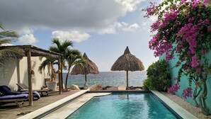 A pool area with lounge chairs, a thatched umbrella, and a view of the sea. - PM78 Boutique Apartments (Willemstad)