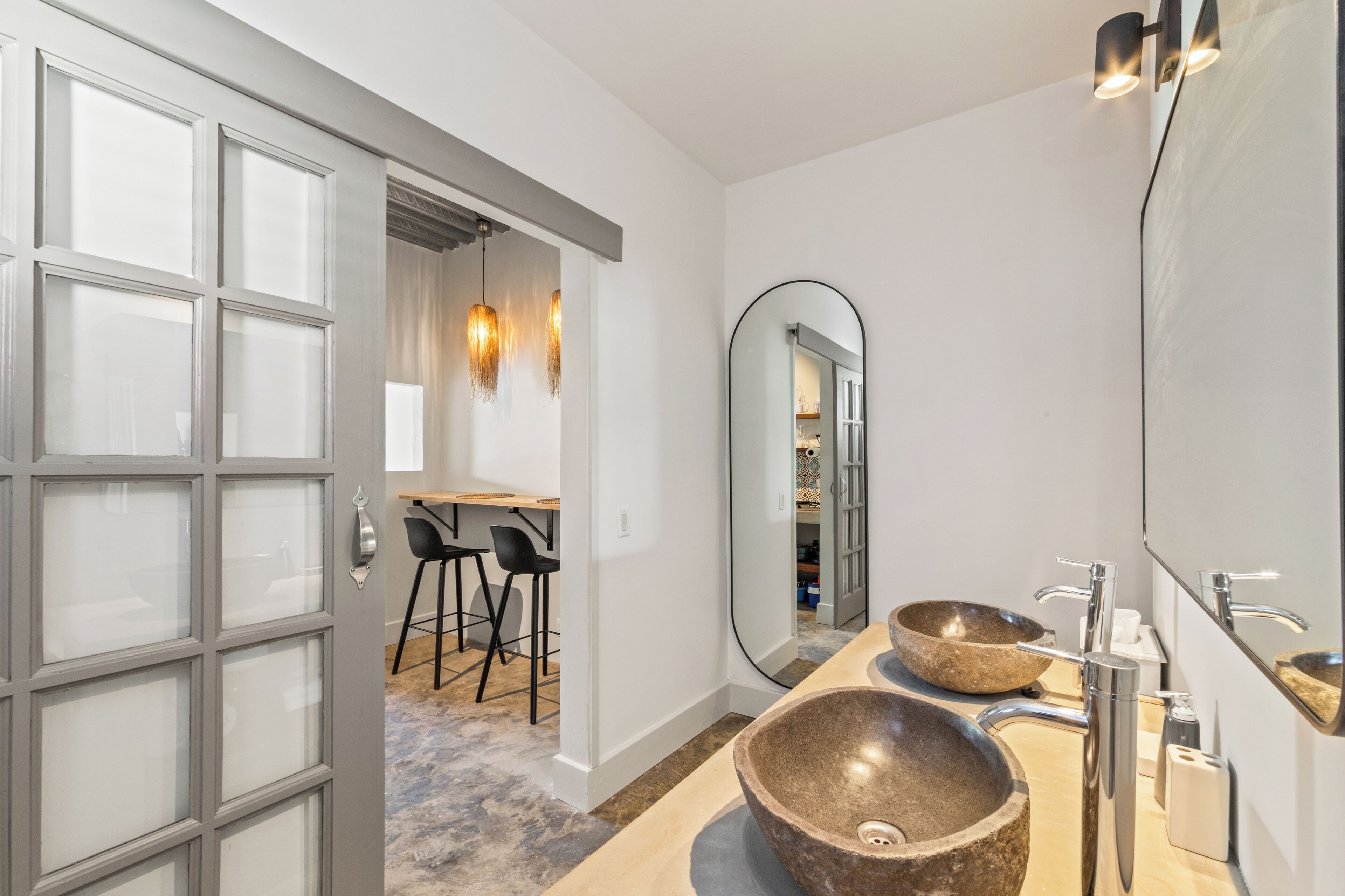 Un baño moderno con dos lavamanos de piedra grandes, un espejo amplio y una puerta que da a un comedor con taburetes de bar.