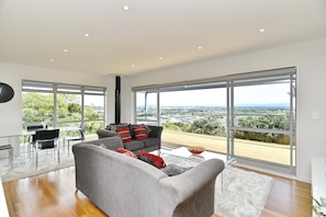 Uma sala de estar espaçosa, com um sofá cinza, almofadas vermelhas e pretas, uma mesa de centro de vidro e vista para o horizonte urbano através de grandes janelas.