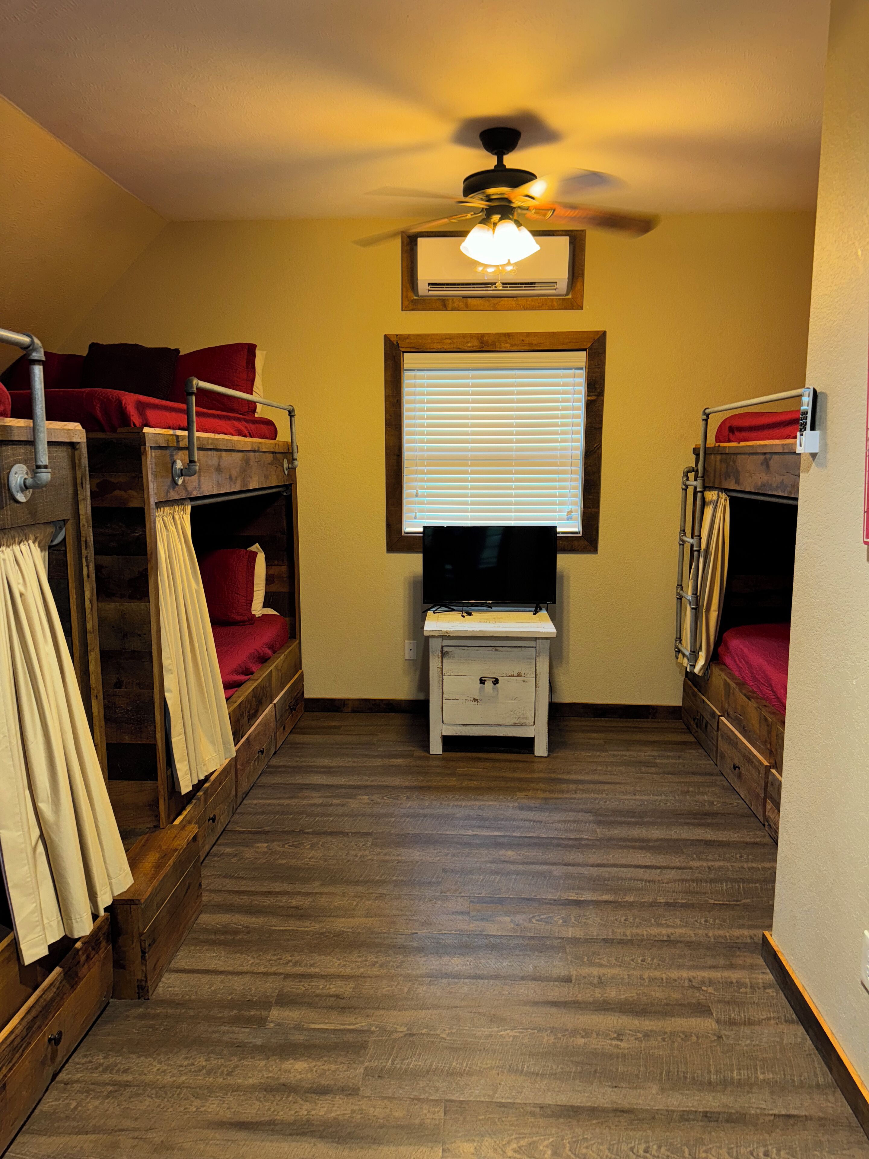 A dormitory room with bunk beds, a small white nightstand, a ceiling fan, and a window with blinds.