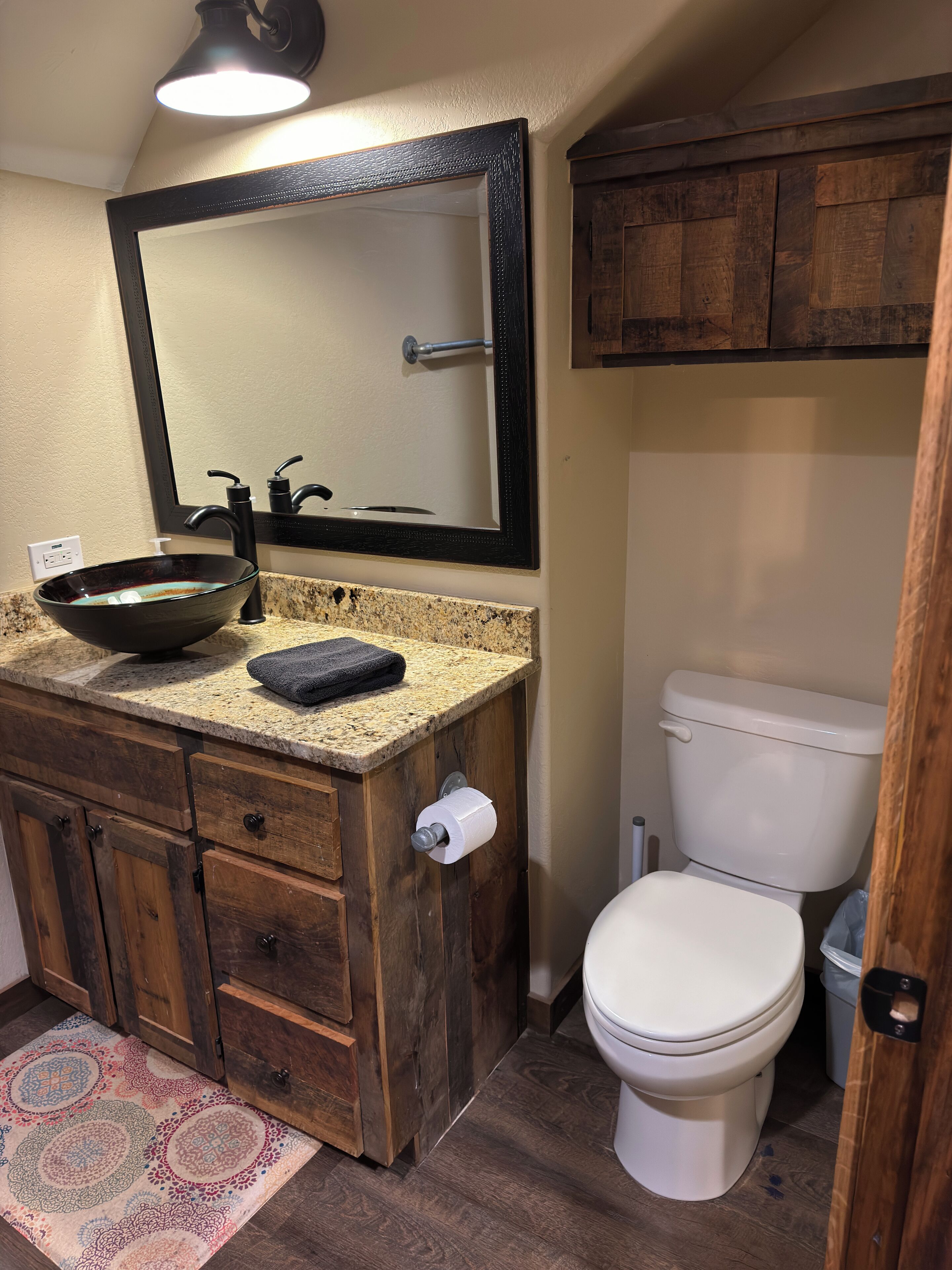 A bathroom with a wooden vanity, a large mirror, a vessel sink, a toilet, and a patterned rug.