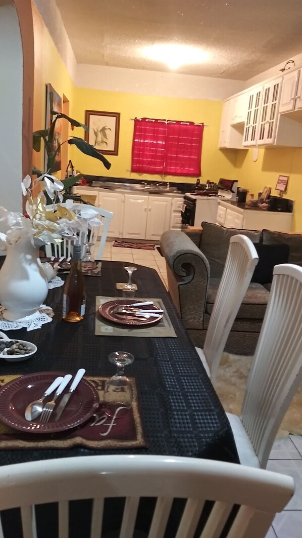 Un comedor con una mesa dispuesta para cuatro, sillas, un florero con flores y una cocina al fondo.