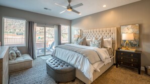 A bedroom with a large bed, a ceiling fan, a nightstand, and a view of the outdoors.