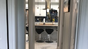 En gang der fører til et køkken med hvide skabe, en marmorbordplade og to sorte barstole.