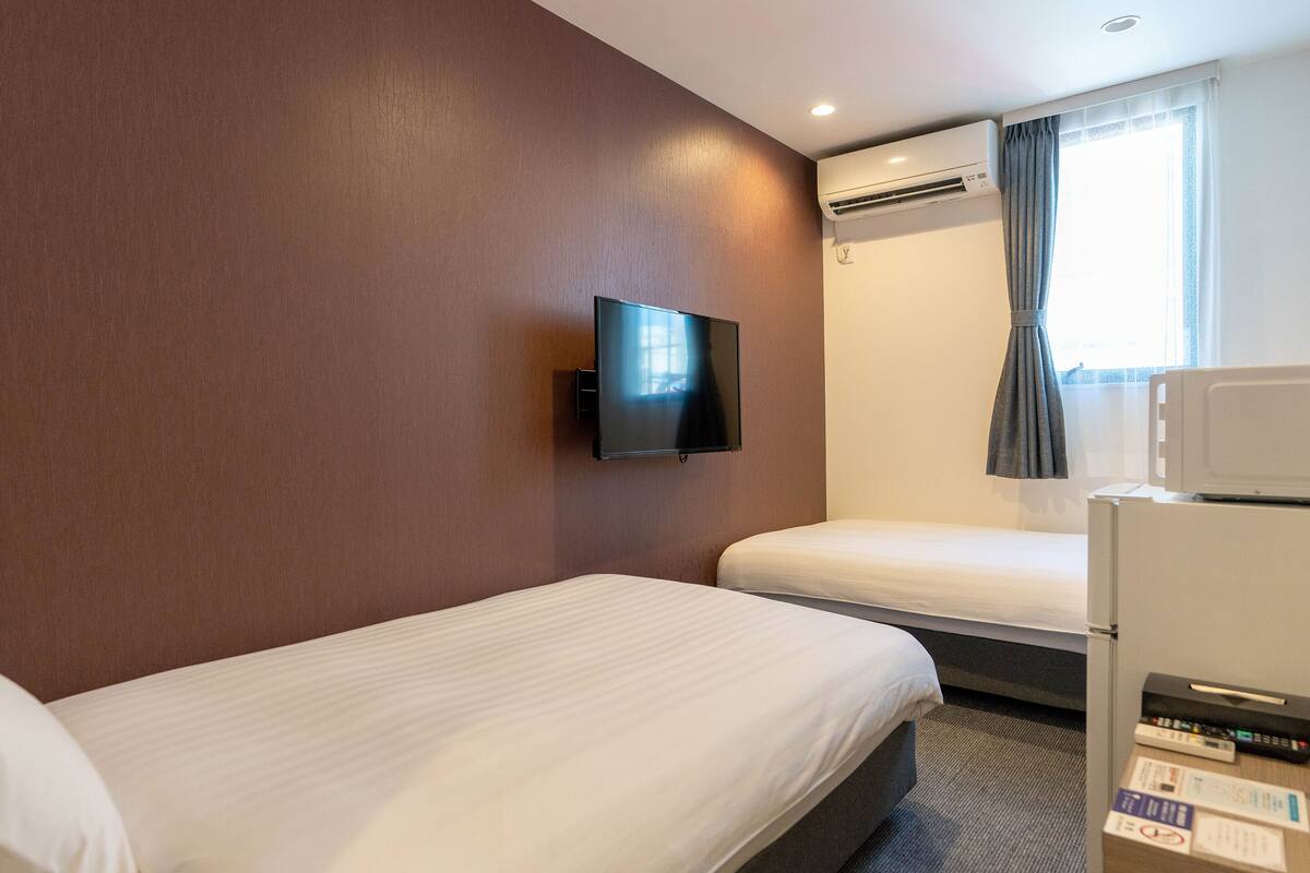 A hotel room with two beds, a flat-screen TV mounted on the wall, an air conditioning unit, a microwave, and a window with curtains.