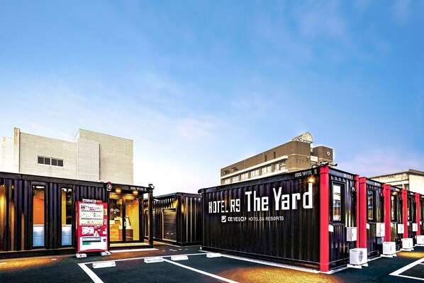 A hotel with a modern design, featuring a prominent 'Hotel R9 The Yard' sign, a vending machine, and a parking lot. - Hotel R9 The Yard Togane (Togane)