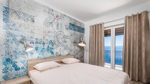Quarto com uma cama grande, mesa de cabeceira, parede com padrão azul e branco, janela com cortinas e vista para o mar.