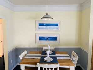 Un comedor con una mesa de madera dispuesta para cuatro personas, sillas blancas y dos cuadros en la pared.