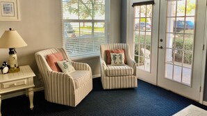 Quarto com duas poltronas listradas, uma pequena mesa com abajur, uma janela grande com persianas e uma porta com vista para o exterior.