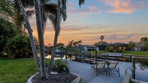 A wooden deck with chairs and a table, overlooking a calm water body and a sunset sky. - Pool Table, Bicycles, Pool & Spa --ESCAPE - Caribbean Getaway - Roelens Vacations (Cape Coral)