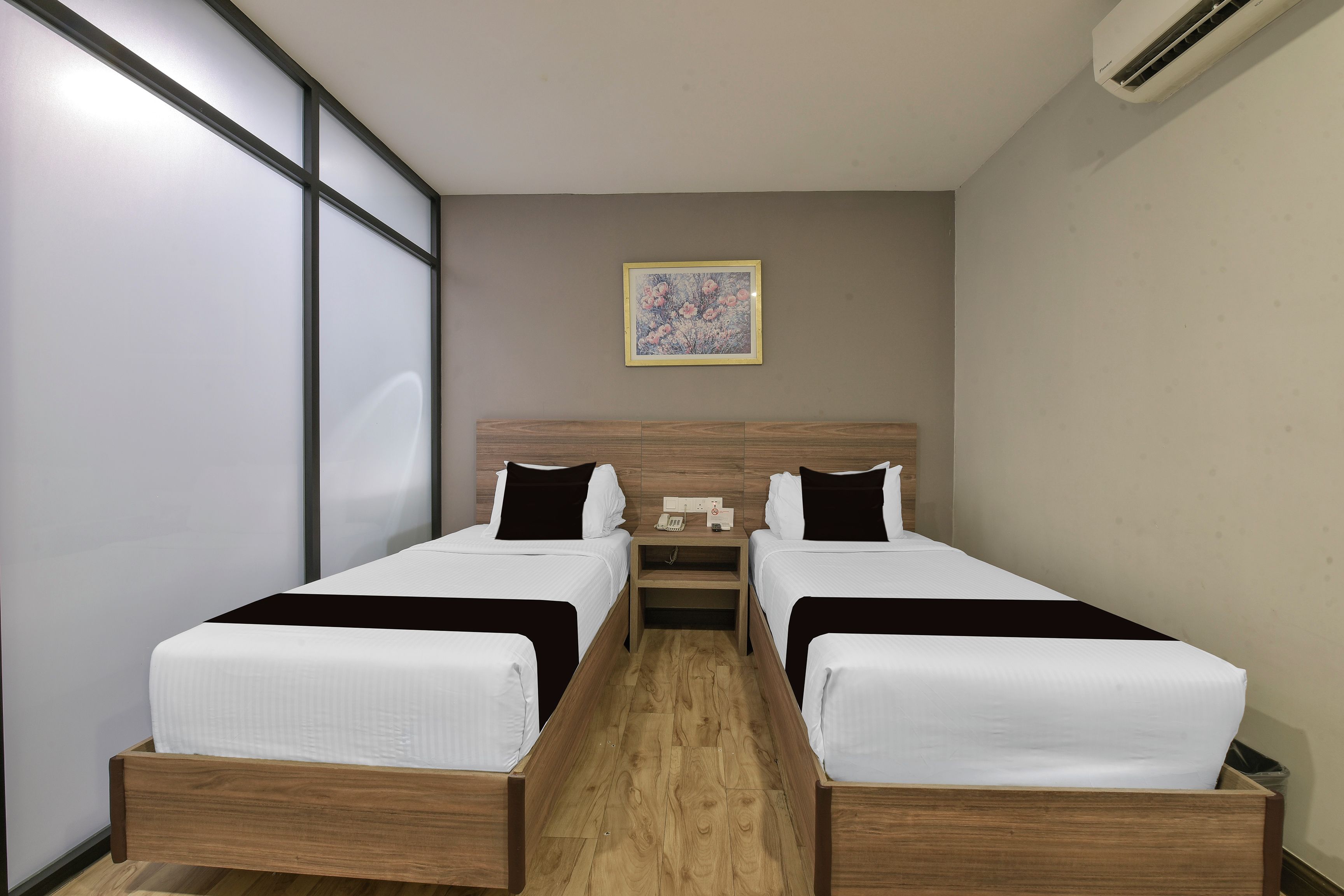 Een hotelkamer met twee éénpersoonsbedden, een houten hoofdbord, een nachtkastje met een telefoon en een klein horloge, en een ingelijst schilderij aan de muur.