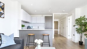 A modern apartment with a kitchen, two bar stools, a grey sofa, and a coffee table with a vase. - One Bedroom Lush Apartment Next to Wembley Stadium (Wembley Park)