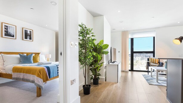 A modern hotel room with a bed, a desk, a potted plant, and a large window offering a city view. - One Bedroom Lush Apartment Next to Wembley Stadium (Wembley Park)