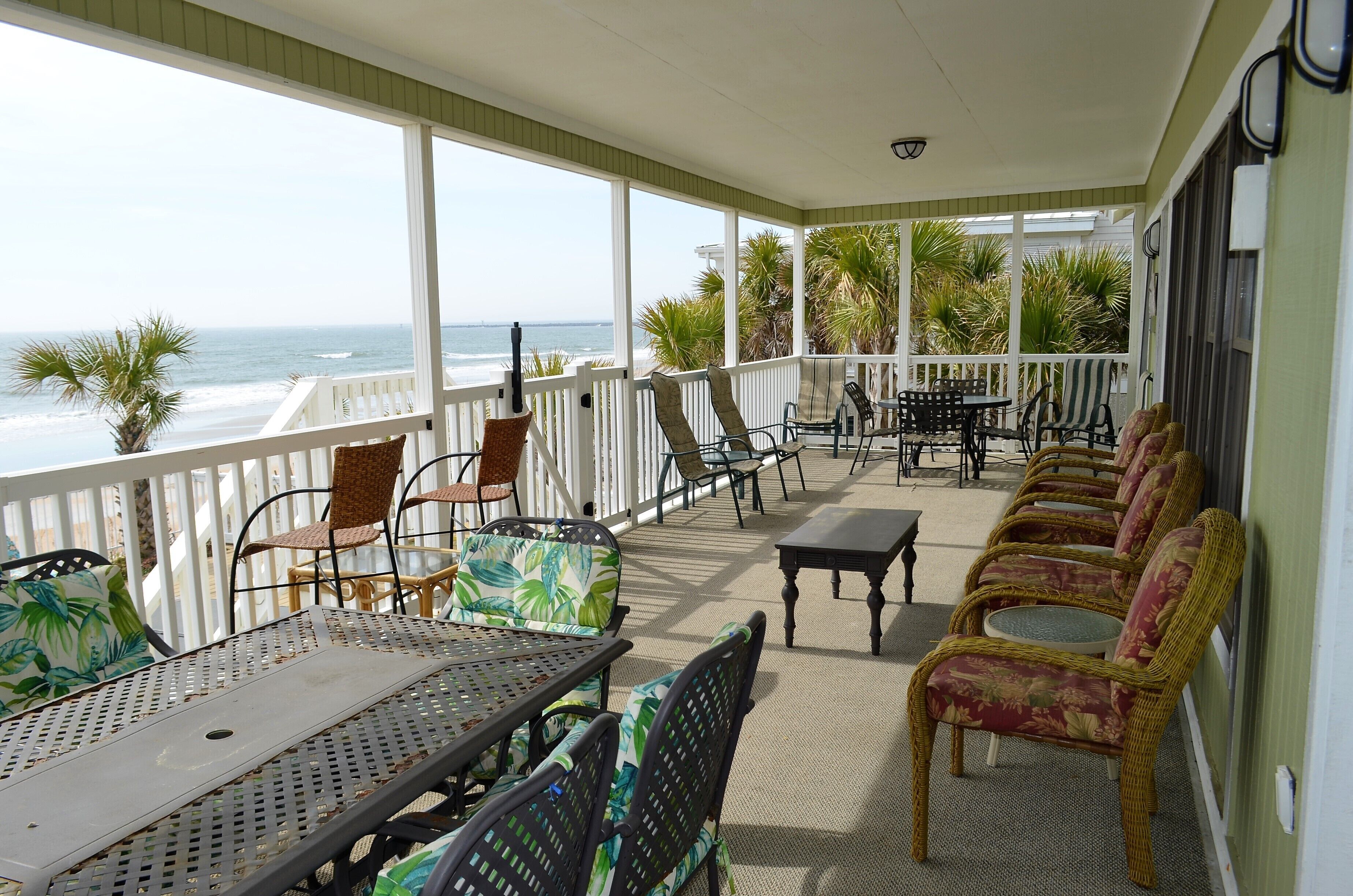 A covered outdoor area with a view of the ocean, featuring wicker chairs, metal tables, and a white railing.