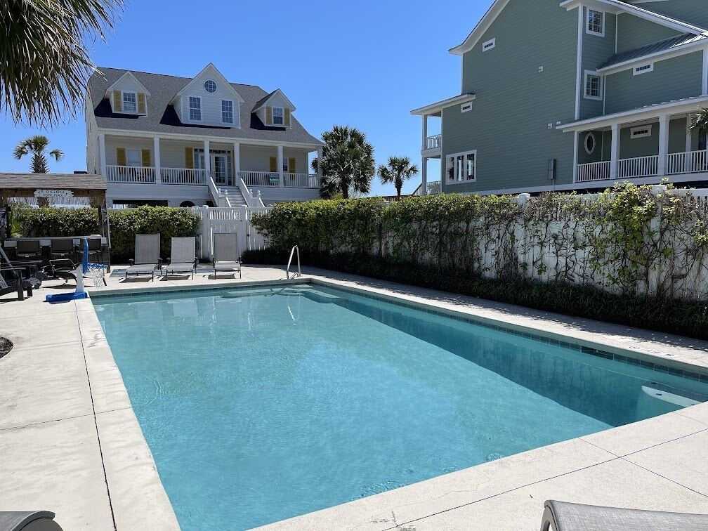 A rectangular swimming pool with a white deck, surrounded by a low hedge and a white fence.