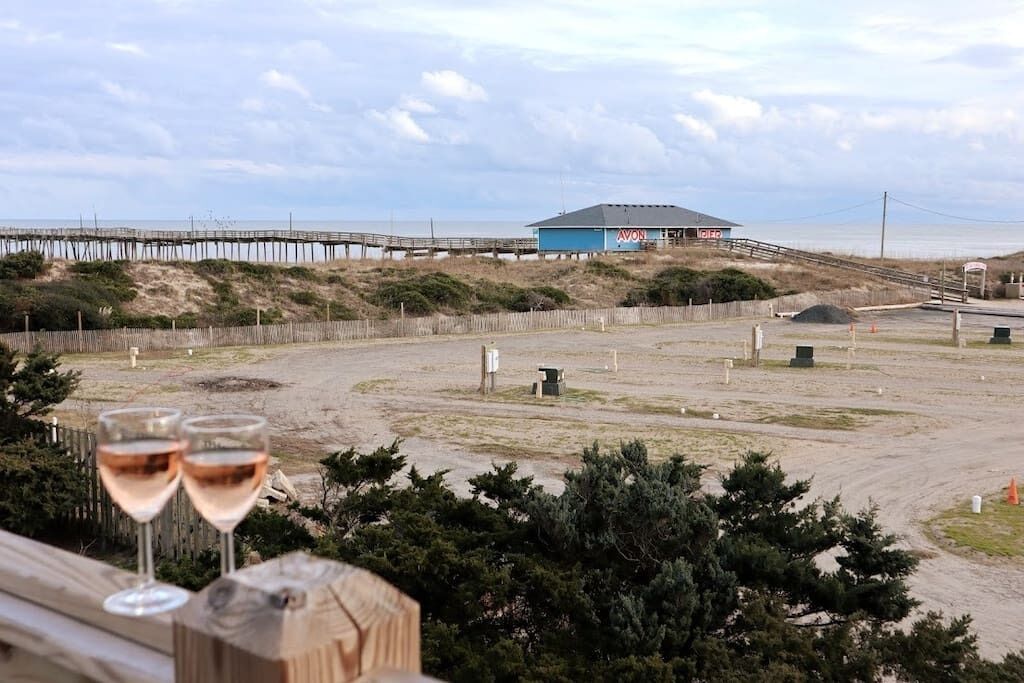 Vista sulla spiaggia con un pontile di legno, due bicchieri di vino e un molo all'orizzonte.