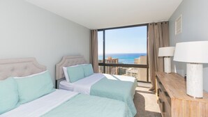 Une chambre d’hôtel avec deux lits, une table de chevet en bois et une grande fenêtre donnant sur la ville et l’océan.