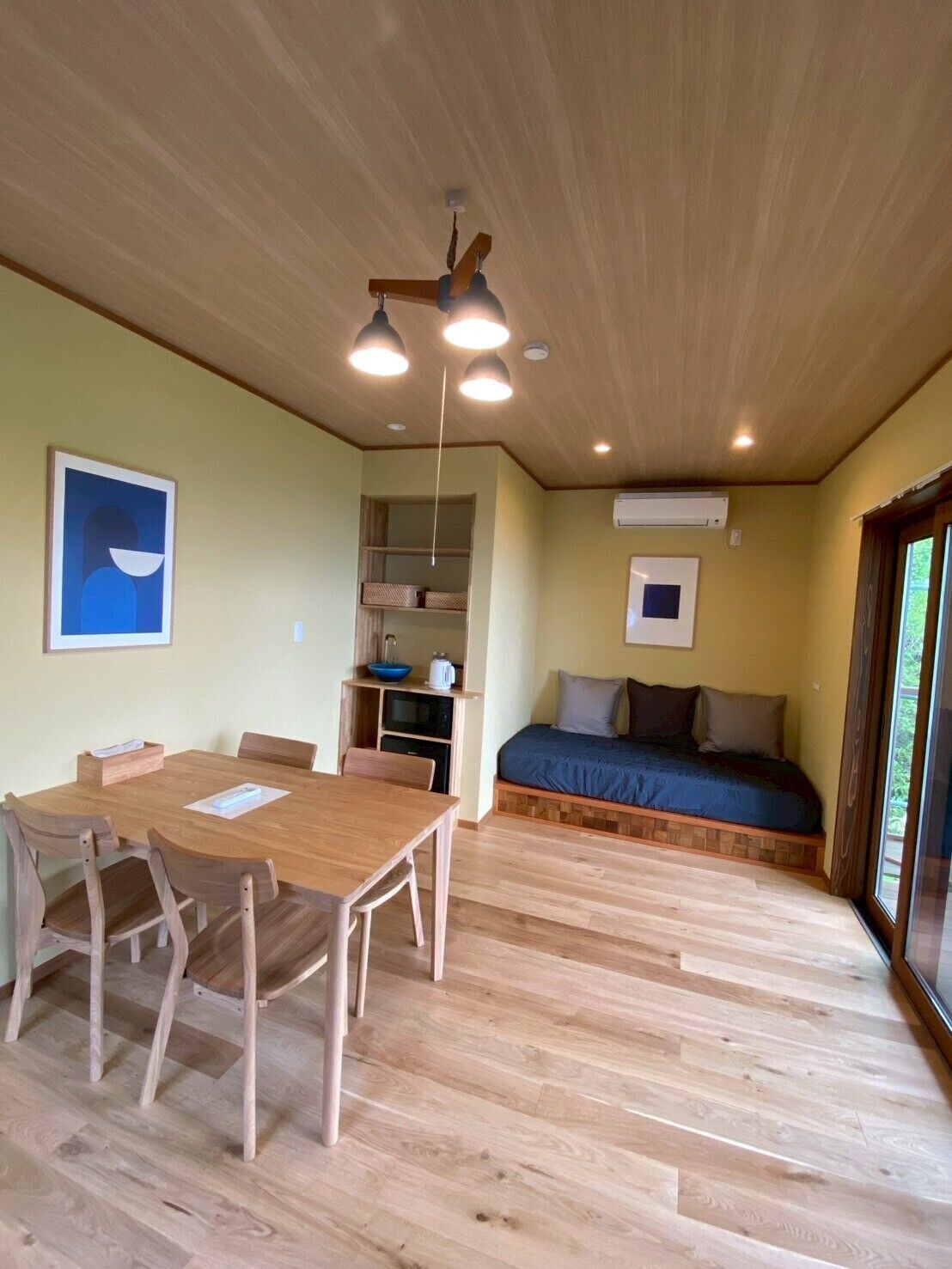 Una stanza con un tavolo e sedie in legno, un letto con materasso blu, una piccola mensola con oggetti e un quadro appeso al muro.