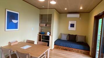 Una stanza con un tavolo e sedie in legno, un letto con materasso blu, una piccola mensola con oggetti e un quadro appeso al muro.