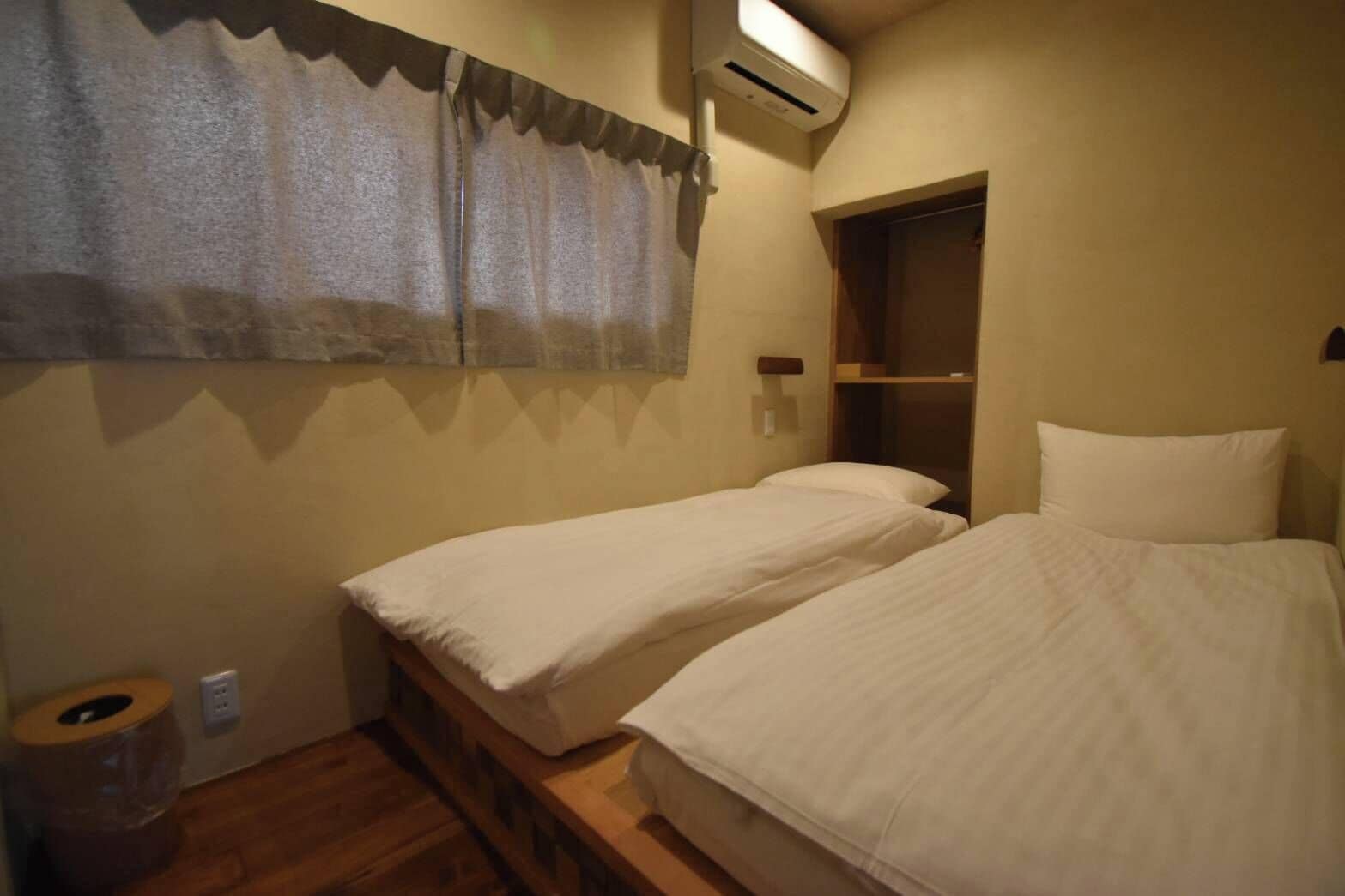 Una habitación pequeña y acogedora con dos camas individuales, suelo de madera, ventana con cortina y armario empotrado.