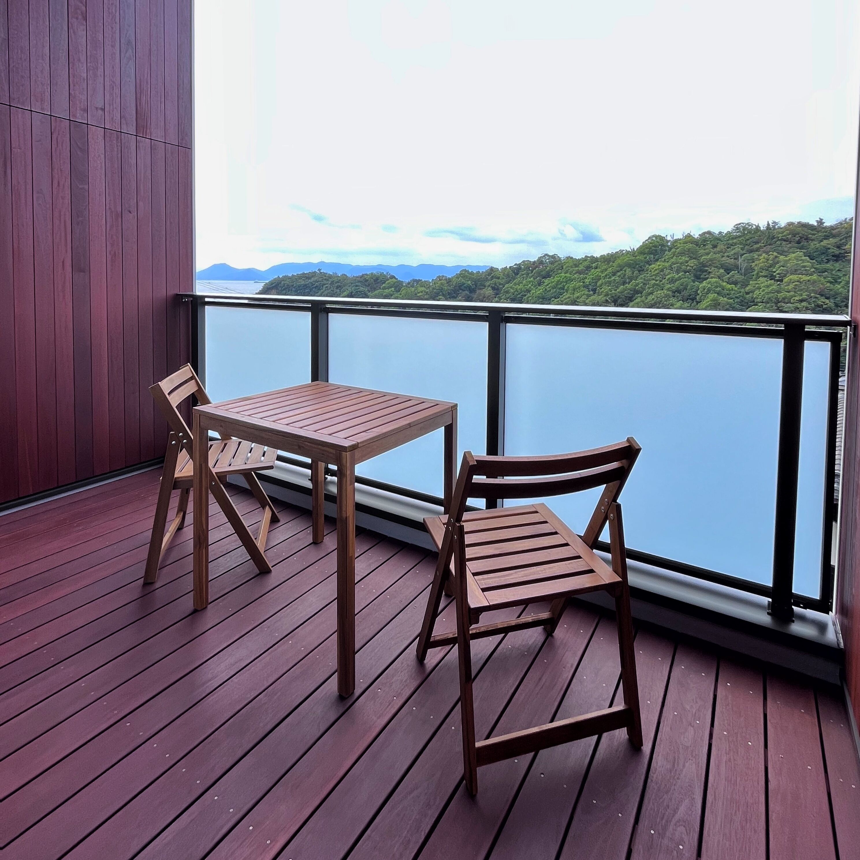 A wooden deck with a table and two chairs, overlooking a body of water and green hills.