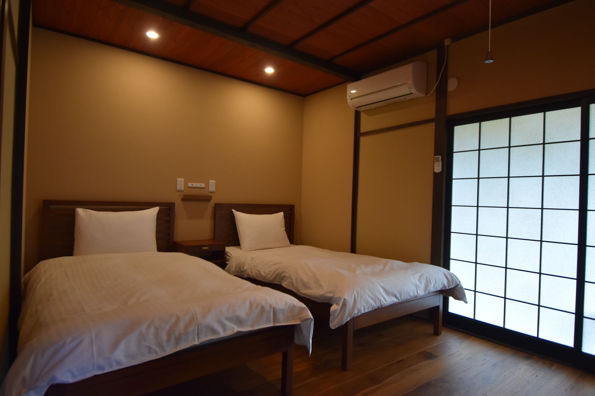 A room with two beds, wooden flooring, a ceiling with recessed lighting, an air conditioning unit, and a sliding glass door with a grid pattern.