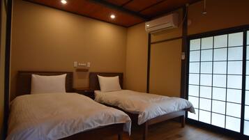 A room with two beds, wooden flooring, a ceiling with recessed lighting, an air conditioning unit, and a sliding glass door with a grid pattern.