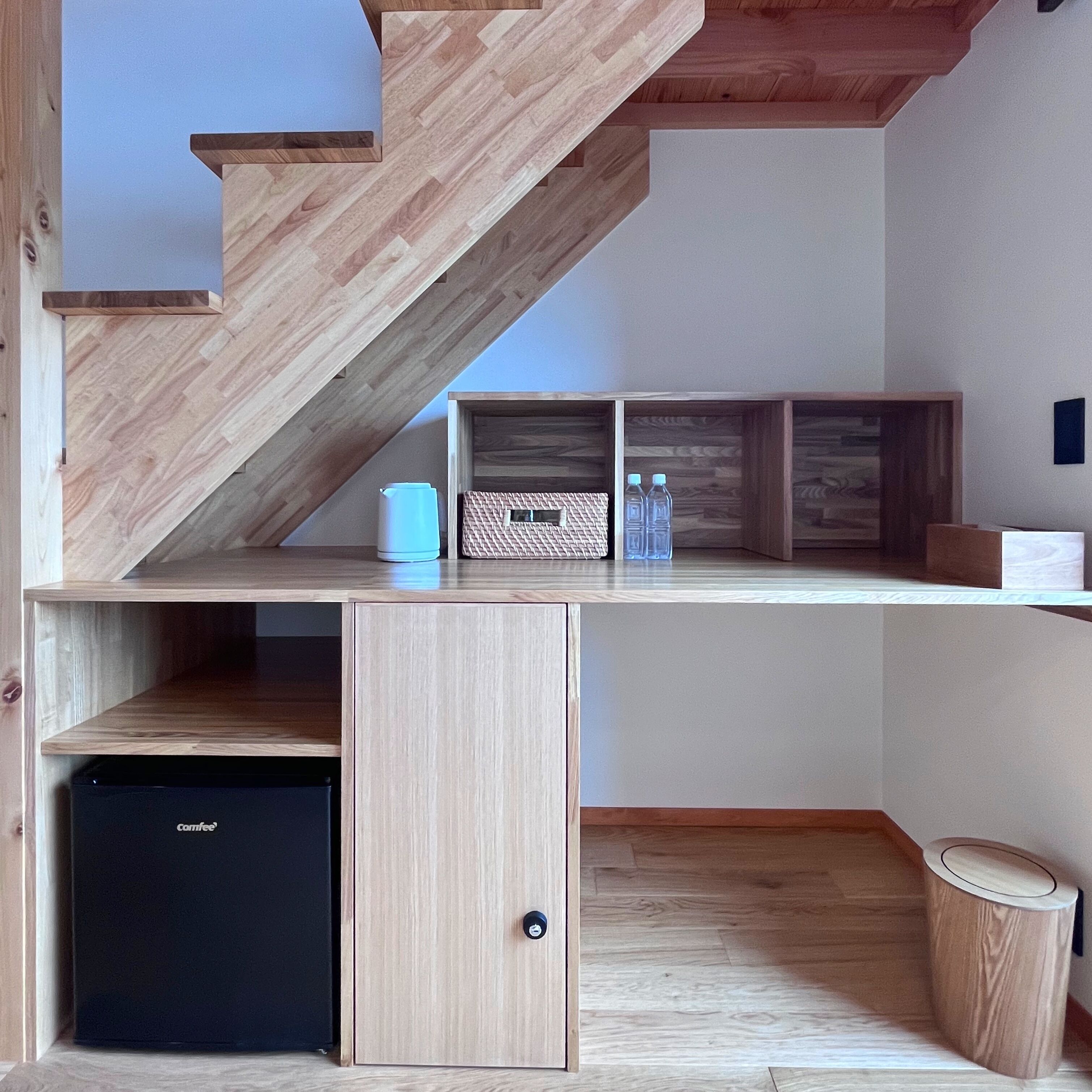 A compact living space with a wooden staircase, built-in shelving, a small black refrigerator, and a trash bin.